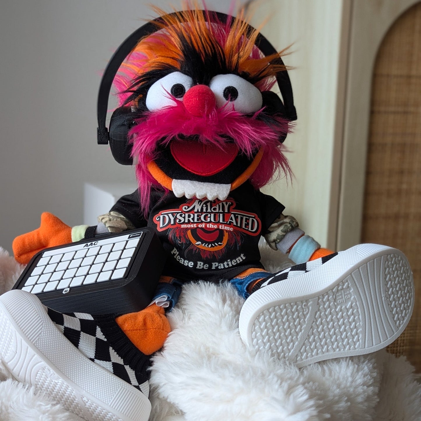 A plush character doll with bright pink, orange, and black hair sits on a white fluffy blanket indoors. The doll wears black headphones, a black graphic T-shirt, and white sneakers, and is holding a small black keyboard.